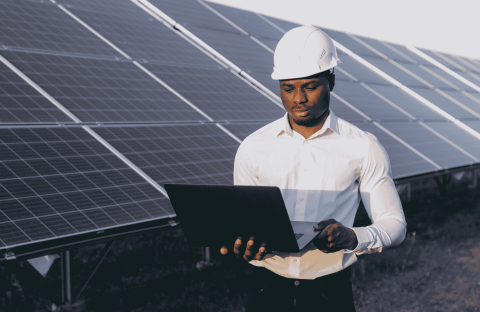 A man wearing a white hard hat and shirt stands outdoors in front of solar panels, holding and working on a laptop to ensure efficient service delivery.