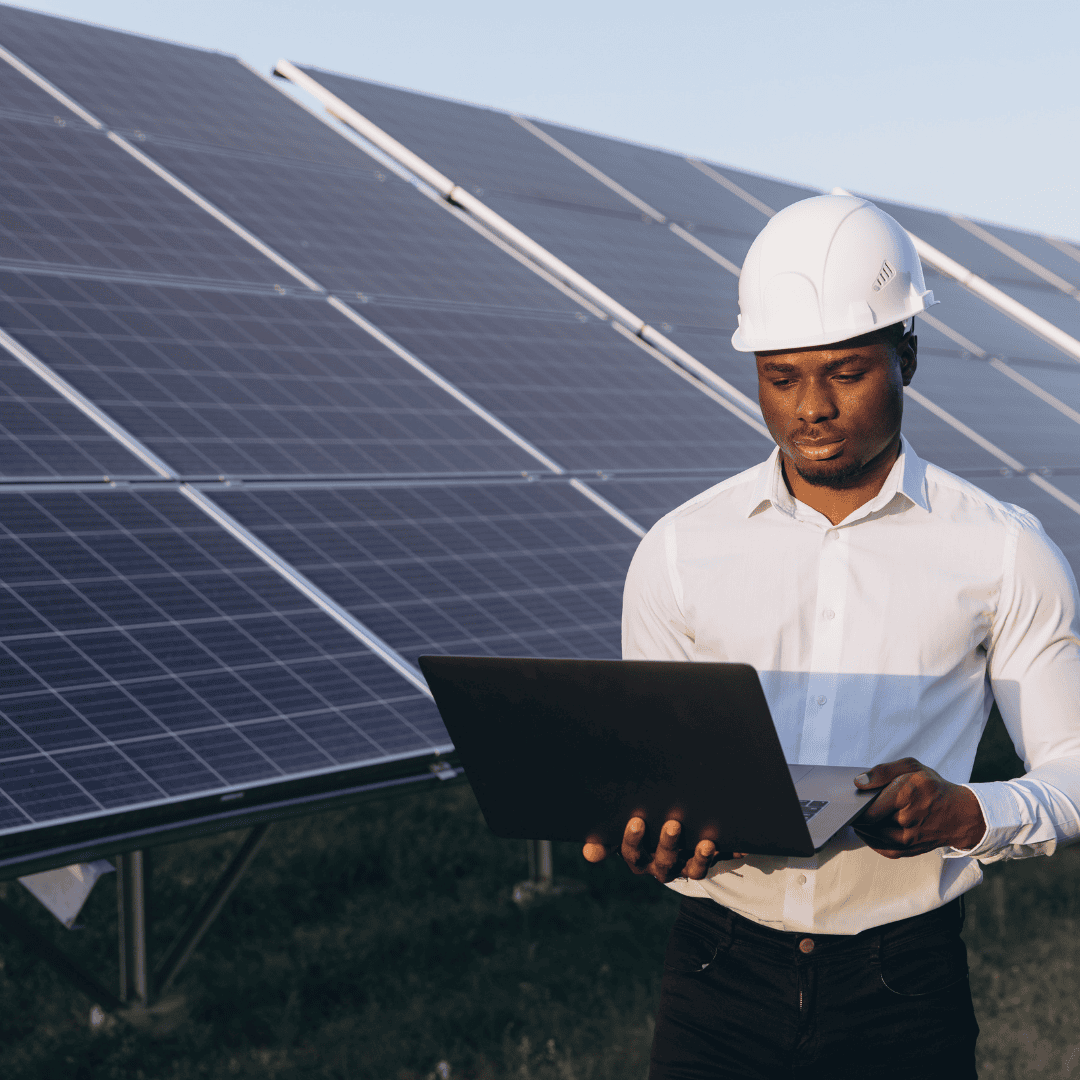 A man wearing a white hard hat and shirt stands outdoors near large solar panels, working on a laptop to monitor service delivery at a solar energy installation site.