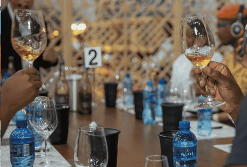 A group of people sit around a table holding up glasses of wine for a toast. The table is set with water bottles, empty wine glasses, and black cups—perhaps a brief escape from daily brainrot. The background features a wooden lattice partition.