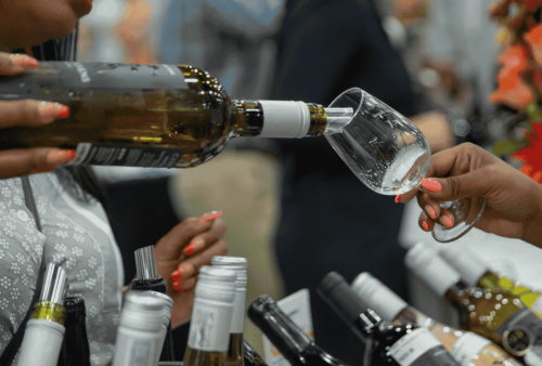 A woman pours white wine from a bottle into a glass held by another person at a wine tasting event, with several other bottles chilling in an ice bucket—perfect for escaping the brainrot of everyday routines.