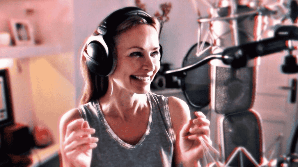 A woman wearing headphones and a sleeveless top smiles while standing in front of a professional microphone in a recording studio.