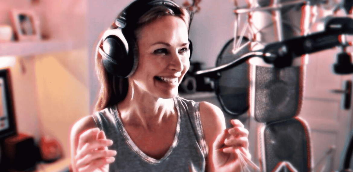 A woman wearing headphones and a sleeveless top smiles while standing in front of a professional microphone in a recording studio.