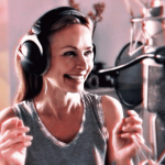 A woman wearing headphones and a sleeveless top smiles while standing in front of a professional microphone in a recording studio.