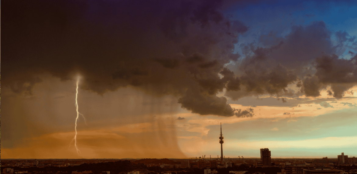 A dramatic cityscape at sunset shows dark storm clouds, a bolt of lightning, and rain falling over part of the city, with orange and blue tones in the sky and a tall communications tower in the distance.
