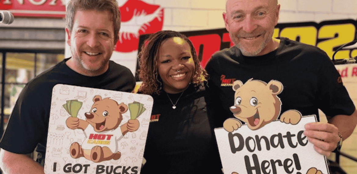 Three smiling adults stand together. Two men hold cartoon bear signs: one reads “I Got Bucks,” the other “Donate Here!” A woman stands between them. All wear black shirts; an Advanced Hair Clinic ad and a radio station logo are visible in the background.