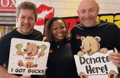 Three smiling adults stand together. Two men hold cartoon bear signs: one reads “I Got Bucks,” the other “Donate Here!” A woman stands between them. All wear black shirts; an Advanced Hair Clinic ad and a radio station logo are visible in the background.
