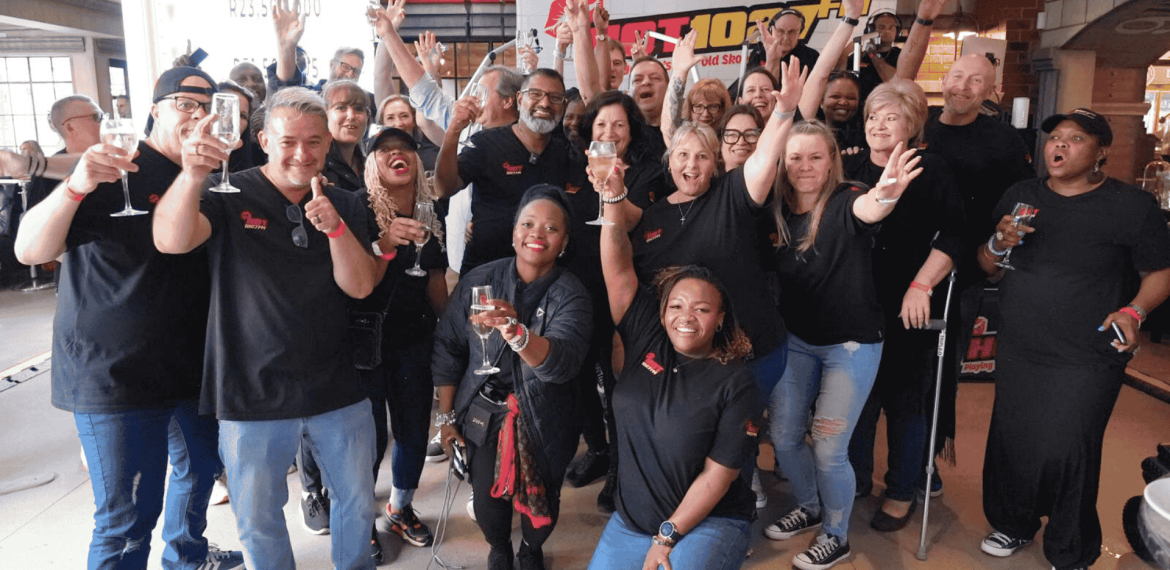 A large, diverse group of people in matching black shirts smiles and celebrates together indoors at Teddython 2025, raising glasses and posing enthusiastically for the camera.