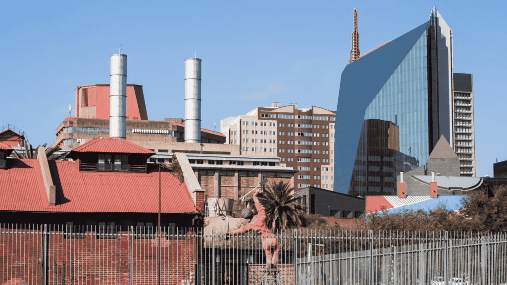 A cityscape with a mix of modern glass buildings and older red-roofed structures, including industrial chimneys, a palm tree in the foreground, and the Olivedale Windmill, all beneath a clear blue sky.