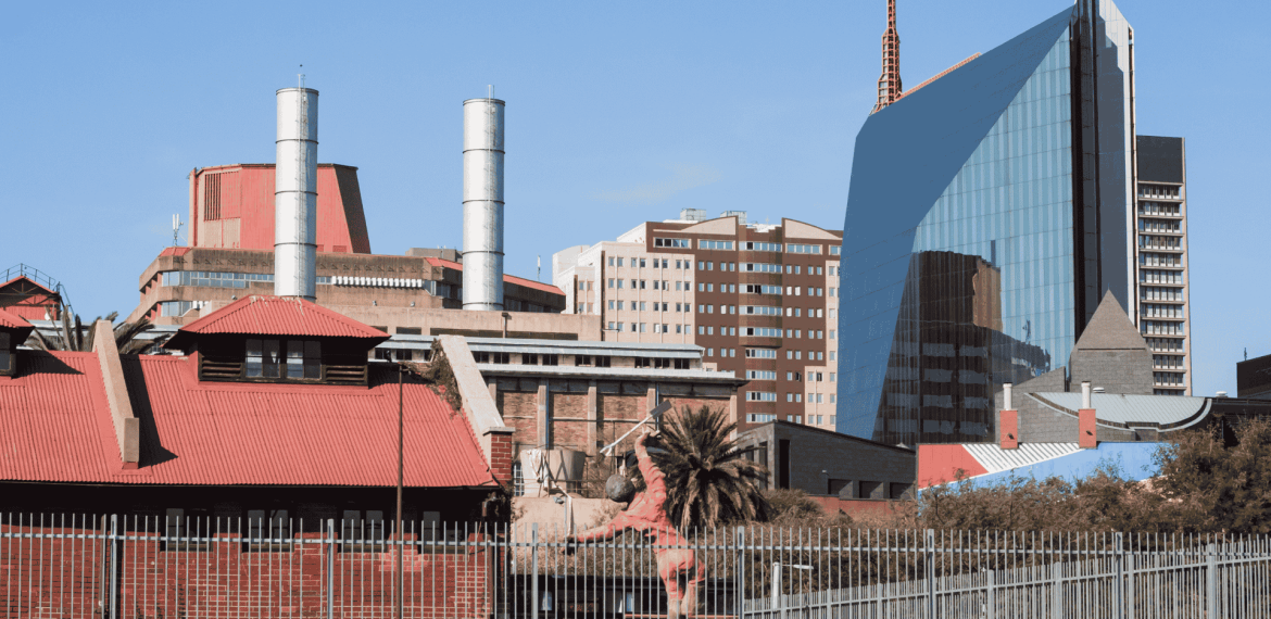 A cityscape with a mix of modern glass buildings and older red-roofed structures, including industrial chimneys, a palm tree in the foreground, and the Olivedale Windmill, all beneath a clear blue sky.