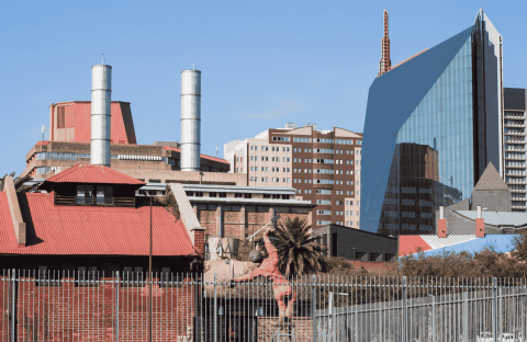 A cityscape with a mix of modern glass buildings and older red-roofed structures, including industrial chimneys, a palm tree in the foreground, and the Olivedale Windmill, all beneath a clear blue sky.