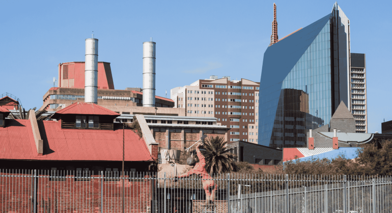 A cityscape with a mix of modern glass buildings and older red-roofed structures, including industrial chimneys, a palm tree in the foreground, and the Olivedale Windmill, all beneath a clear blue sky.