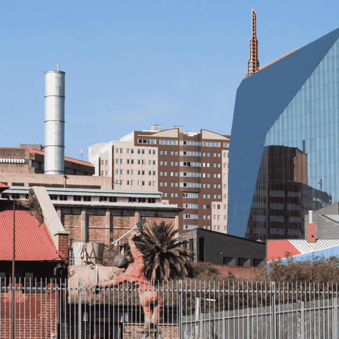 Cityscape with modern and older buildings, including a glass structure, industrial elements, and the Olivedale Windmill. In the foreground, a large pink statue climbs a fence near a palm tree under a clear blue sky.