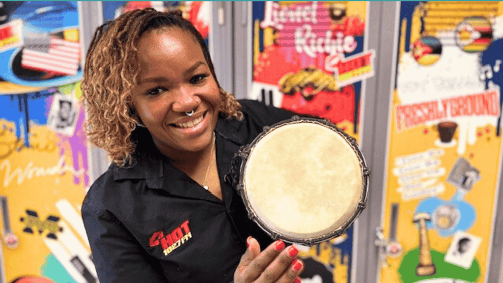 A smiling woman in a black shirt holds a small hand drum towards the camera, with colorful, music-themed posters from Olivedale Windmill displayed on the wall behind her.