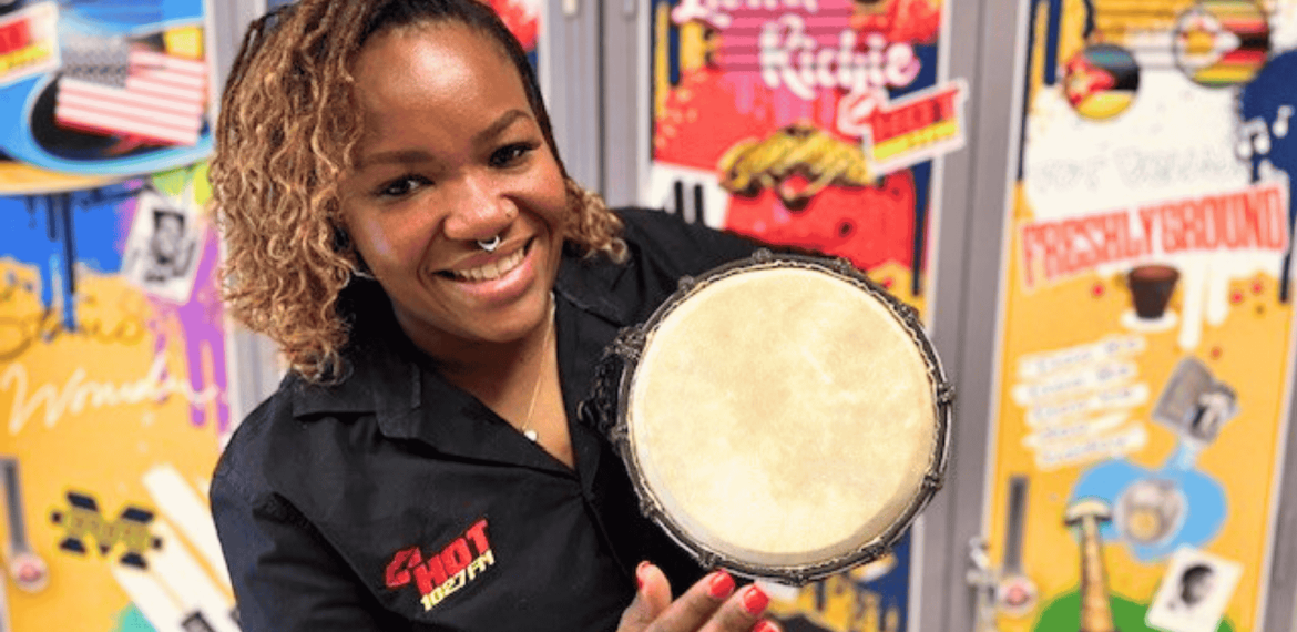A smiling woman in a black shirt holds a small hand drum towards the camera, with colorful, music-themed posters from Olivedale Windmill displayed on the wall behind her.