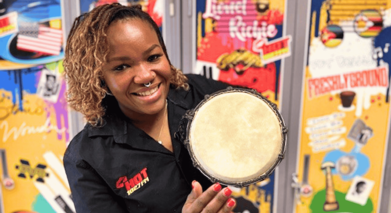 A smiling woman in a black shirt holds a small hand drum towards the camera, with colorful, music-themed posters from Olivedale Windmill displayed on the wall behind her.