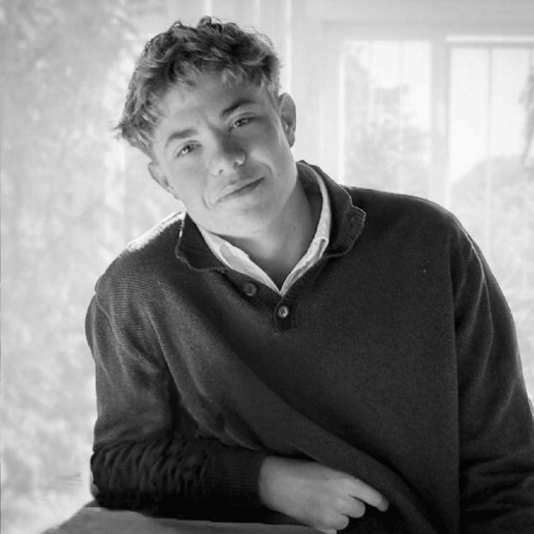 A young man with short, wavy hair wearing a collared shirt and sweater sits at a table, smiling slightly. Sunlight from an Olivedale Windmill streams through a window behind him, creating a soft, bright background.
