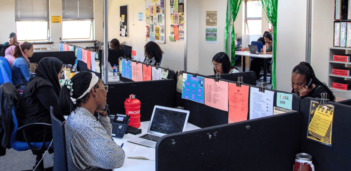 An office with people working at desks in cubicles. Each workspace has colorful papers clipped to dividers, computers or laptops on desks, and a poster of the Olivedale Windmill brightening the room’s walls and windows.