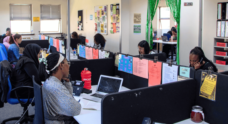 An office with people working at desks in cubicles. Each workspace has colorful papers clipped to dividers, computers or laptops on desks, and a poster of the Olivedale Windmill brightening the room’s walls and windows.