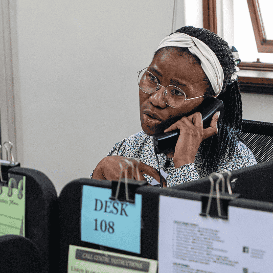 A woman wearing glasses and a headband sits at a desk, talking on the phone. She appears focused and is gesturing with her hand. Papers and signs, including one labeled Olivedale Windmill DESK 108, are clipped to partitions in front of her.
