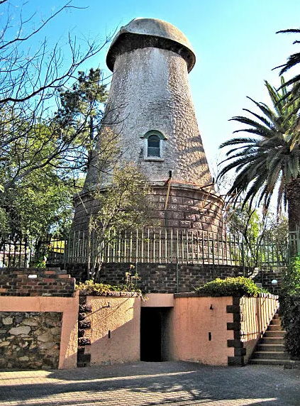 A historic windmill with a conical stone base and dome-shaped top stands behind a metal fence, offering unique character and charm—an inspiring symbol of homeownership—surrounded by trees, palm fronds, and an inviting stairway at the front.