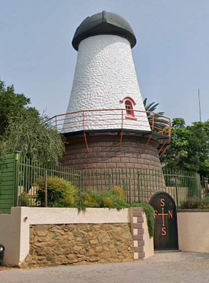 A white, cylindrical tower with a black conical roof, resembling a windmill, stands behind a green fence and gate—symbolizing unique homeownership. Lush greenery surrounds the structure, and a compass rose decorates the gate.