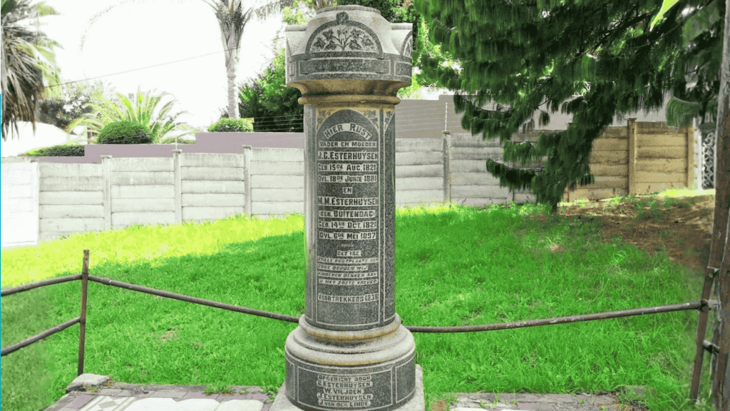 A tall cylindrical stone memorial stands on a lawn in the Magaliesberg area, engraved with multiple inscriptions and names. It is surrounded by a low metal railing, with trees, grass, and a fence in the background.
