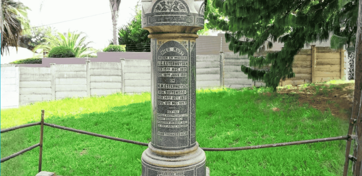A tall cylindrical stone memorial stands on a lawn in the Magaliesberg area, engraved with multiple inscriptions and names. It is surrounded by a low metal railing, with trees, grass, and a fence in the background.