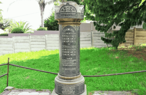 A tall cylindrical stone memorial stands on a lawn in the Magaliesberg area, engraved with multiple inscriptions and names. It is surrounded by a low metal railing, with trees, grass, and a fence in the background.