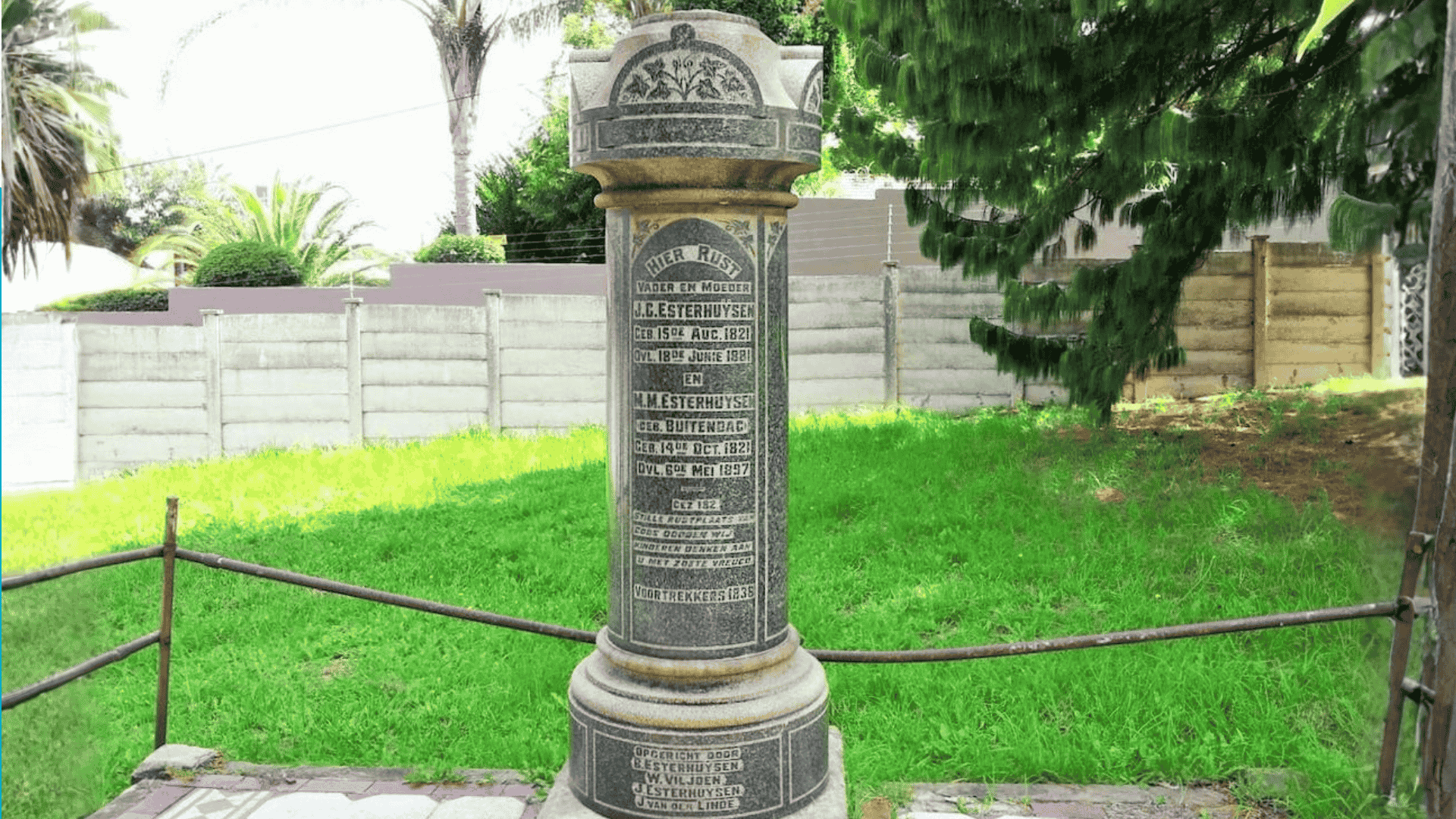A tall cylindrical stone memorial stands on a lawn in the Magaliesberg area, engraved with multiple inscriptions and names. It is surrounded by a low metal railing, with trees, grass, and a fence in the background.