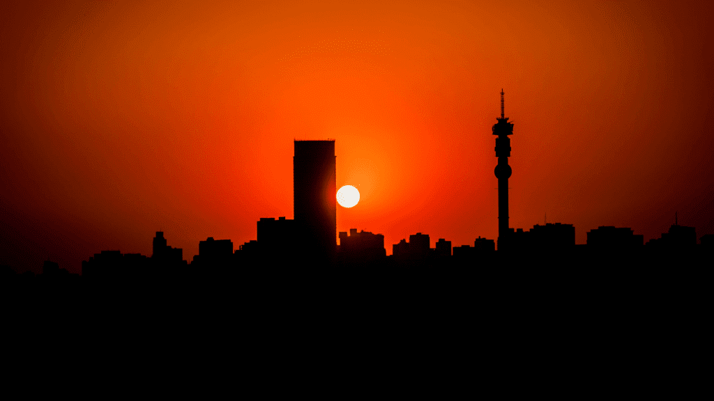 Silhouette of a city skyline at sunset, with the sun partially obscured by a tall building. The sky features a vibrant orange and red gradient reminiscent of Magaliesberg evenings, and a tower with antennas stands on the right side of the image.