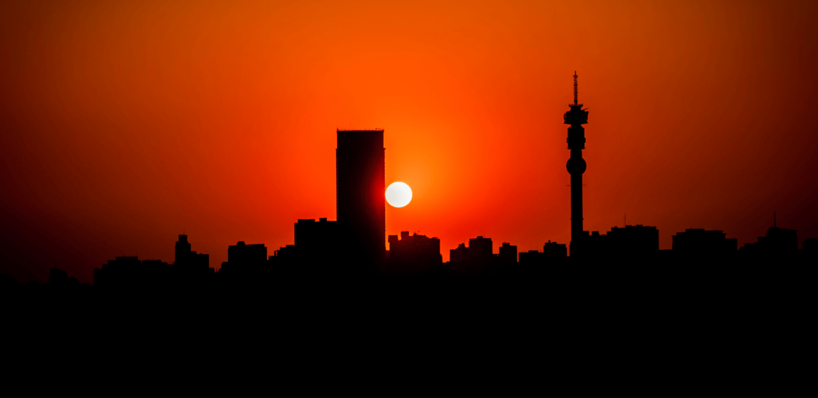 Silhouette of a city skyline at sunset, with the sun partially obscured by a tall building. The sky features a vibrant orange and red gradient reminiscent of Magaliesberg evenings, and a tower with antennas stands on the right side of the image.