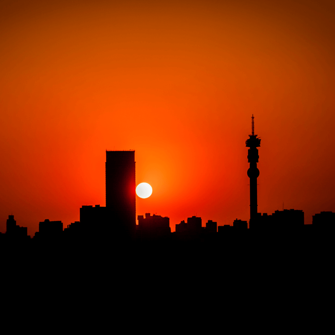 Silhouette of a city skyline at sunset, with the sun partially hidden behind a tall building and an orange-red sky. A tall tower stands out among the buildings, with the distant outline of Magaliesberg visible on the horizon.