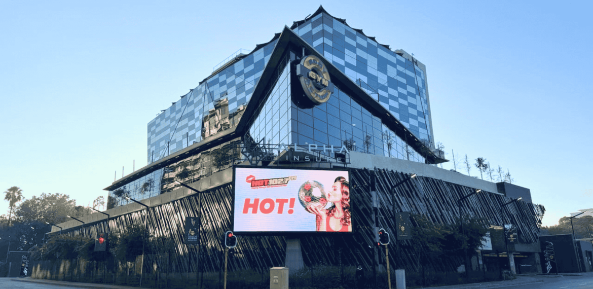 A modern, glass-fronted Hard Rock Hotel building with a large digital billboard displaying an ad featuring Sharon Corr holding a burger and the word HOT! at a city intersection.