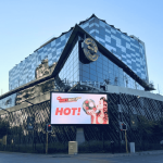 A modern, glass-fronted Hard Rock Hotel building with a large digital billboard displaying an ad featuring Sharon Corr holding a burger and the word HOT! at a city intersection.