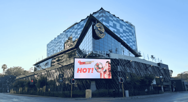 A modern, glass-fronted Hard Rock Hotel building with a large digital billboard displaying an ad featuring Sharon Corr holding a burger and the word HOT! at a city intersection.