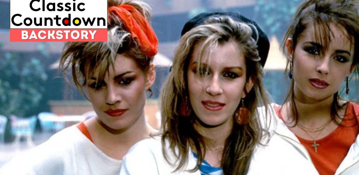 Three women with 1980s hairstyles and makeup stand close together, looking at the camera. A Classic Countdown Backstory logo and a "Love is in the Air" vibe set the scene, while the blurred background hints at a lively outdoor setting.