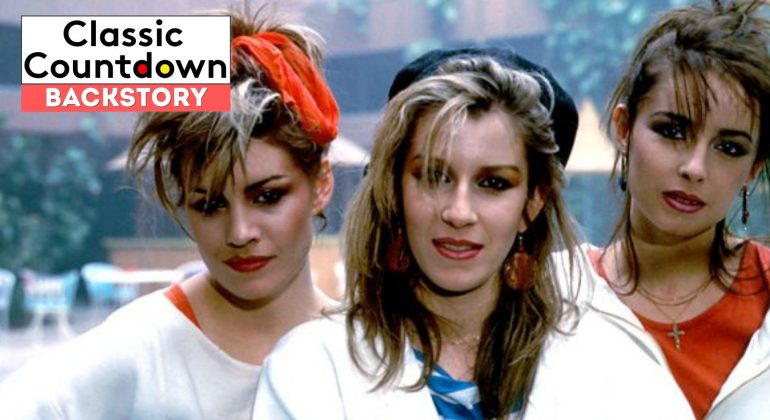 Three women with 1980s hairstyles and makeup stand close together, looking at the camera. A Classic Countdown Backstory logo and a "Love is in the Air" vibe set the scene, while the blurred background hints at a lively outdoor setting.