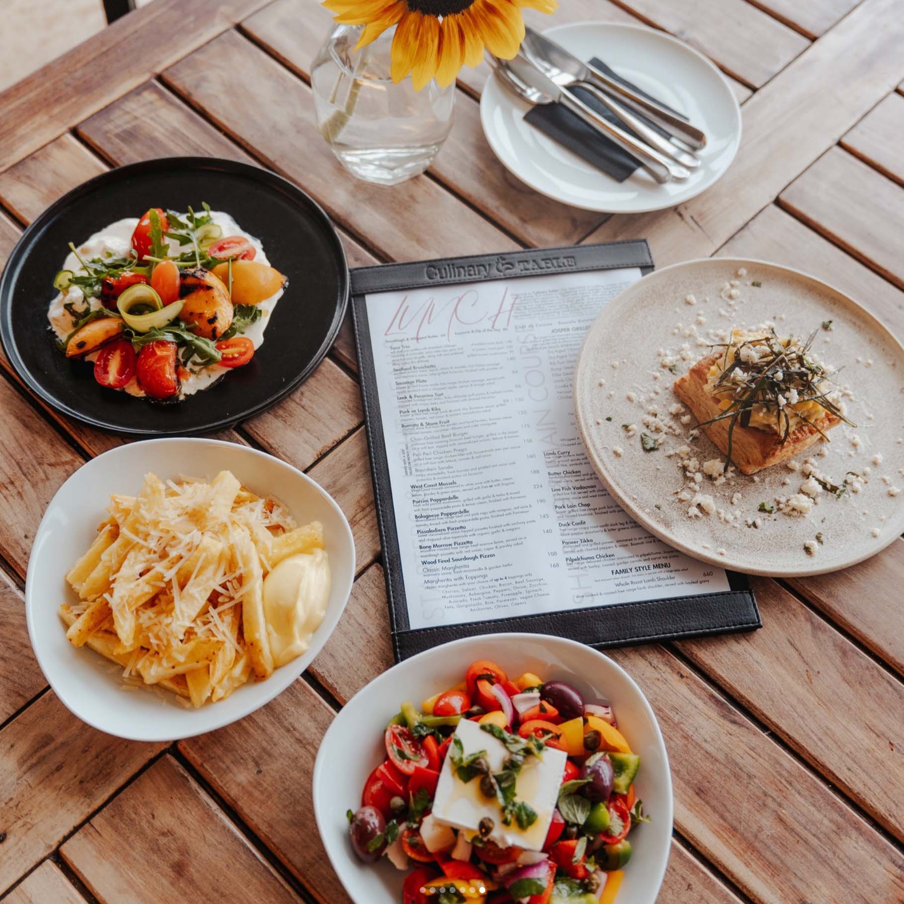 Four plates of assorted dishes, perfect for Heritage Day, including pasta, salad, and an appetizer, surround a restaurant menu on a wooden table. A vase with a sunflower and a set of cutlery are in the background.