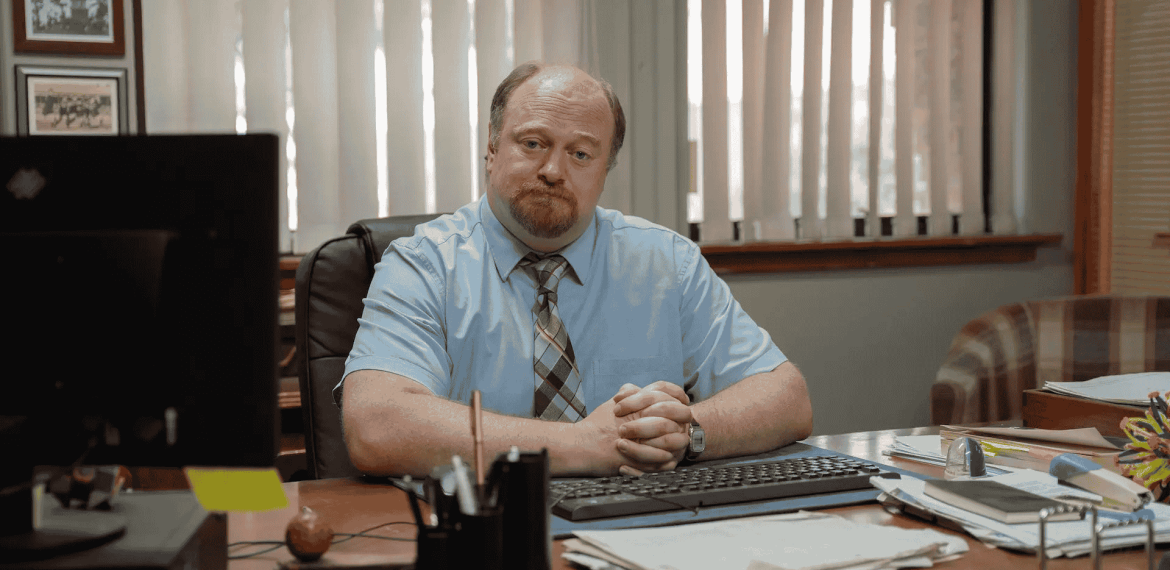 A man with a beard and mustache, wearing a light blue shirt and striped tie, sits at his desk with hands folded. He faces the camera in an office with window blinds, various papers, and notes about road closures on the desk.