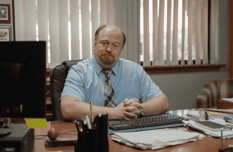 A man with a beard and mustache, wearing a light blue shirt and striped tie, sits at his desk with hands folded. He faces the camera in an office with window blinds, various papers, and notes about road closures on the desk.