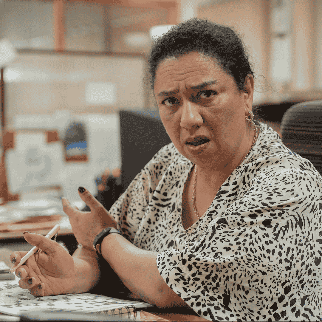 A woman with curly hair, wearing a black-and-white patterned blouse, sits at a desk holding a pen and looks confused or frustrated, gesturing with her hand—perhaps sorting through office papers about unexpected road closures.