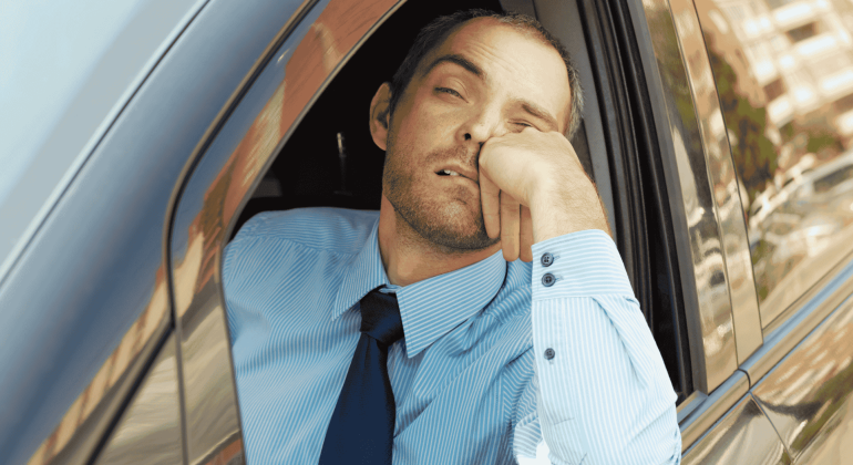 A man in a blue dress shirt and tie rests his head on his hand, looking tired and bored while sitting in a car with the window down, perhaps contemplating new business opportunities.