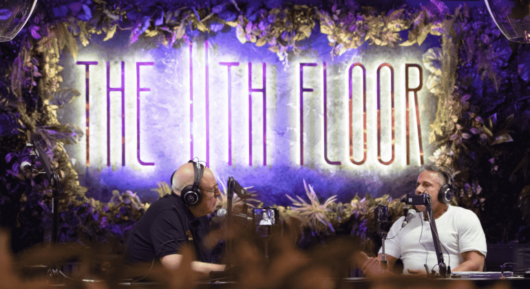 Two men wearing headphones and sitting at microphones discuss business opportunities in front of a sign that reads THE 11TH FLOOR, surrounded by decorative greenery and dramatic lighting.