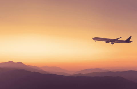 A commercial airplane flies above mountain silhouettes at sunset, with an orange and yellow sky in the background, symbolizing new business opportunities on the horizon.