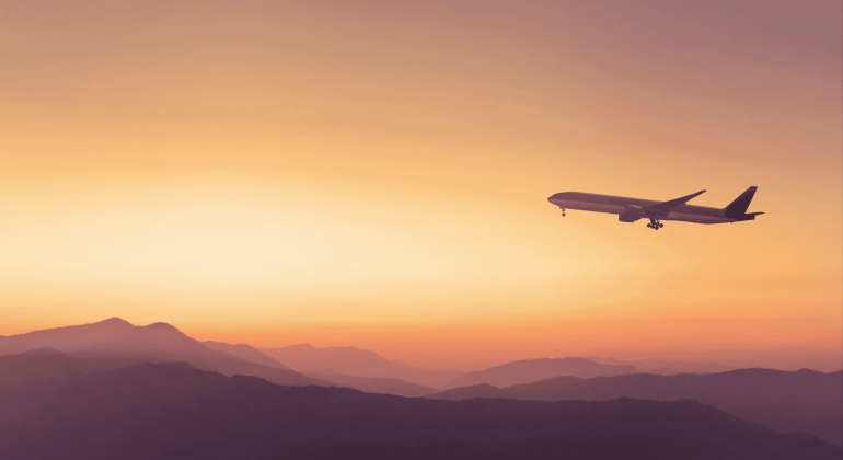 A commercial airplane flies above mountain silhouettes at sunset, with an orange and yellow sky in the background, symbolizing new business opportunities on the horizon.