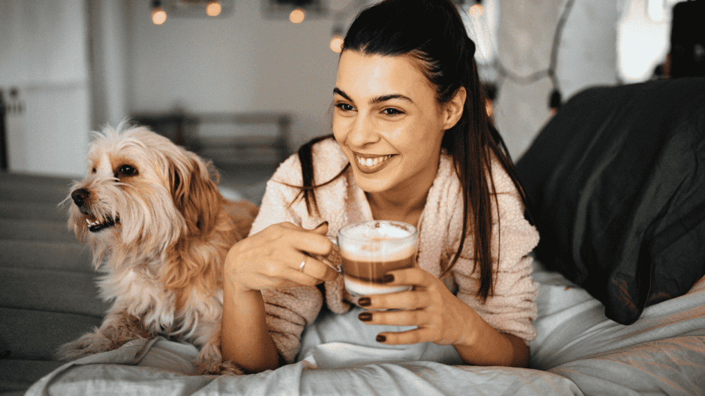 A smiling woman in a bathrobe sits on a bed, holding a cup of coffee, with a fluffy dog beside her. Warm indoor lighting creates a cozy, relaxed atmosphere—an inviting setting to unwind or explore new business opportunities from home.