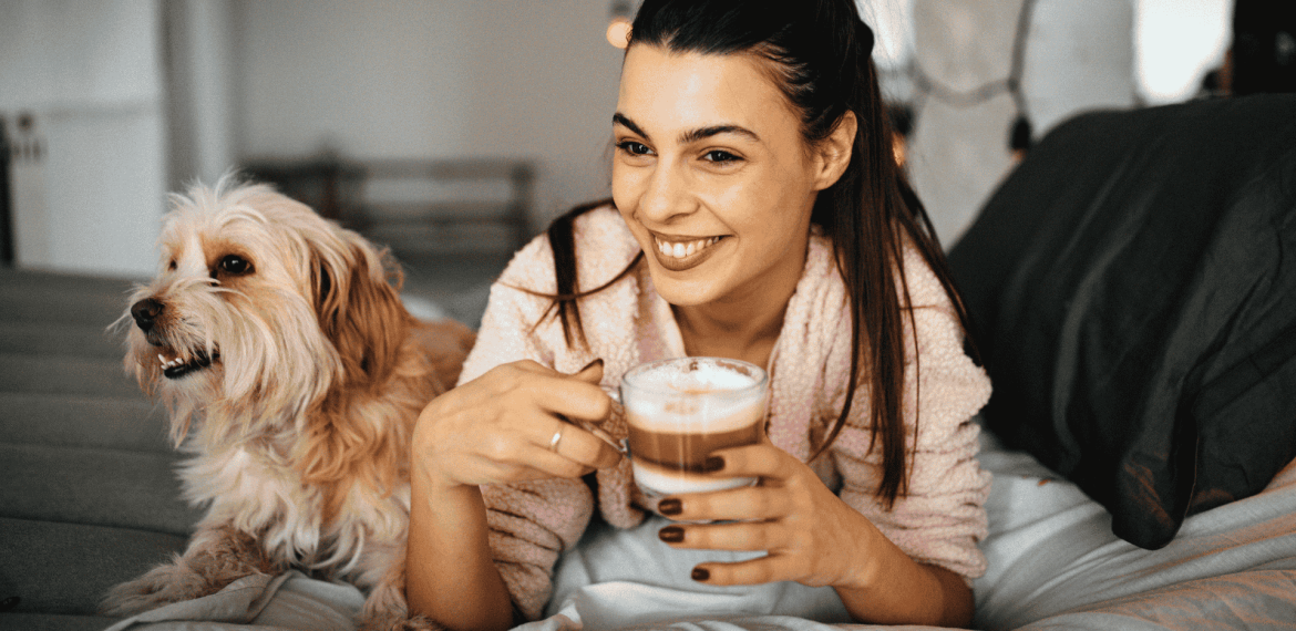 A smiling woman in a bathrobe sits on a bed, holding a cup of coffee, with a fluffy dog beside her. Warm indoor lighting creates a cozy, relaxed atmosphere—an inviting setting to unwind or explore new business opportunities from home.