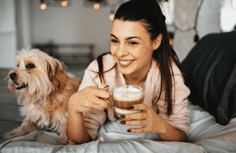 A smiling woman in a bathrobe sits on a bed, holding a cup of coffee, with a fluffy dog beside her. Warm indoor lighting creates a cozy, relaxed atmosphere—an inviting setting to unwind or explore new business opportunities from home.