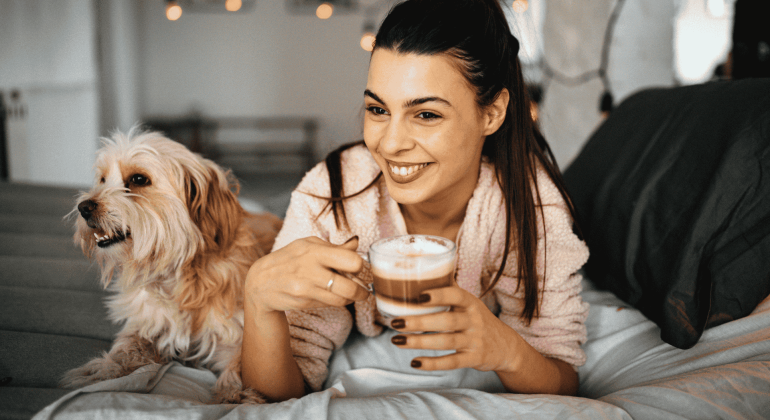 A smiling woman in a bathrobe sits on a bed, holding a cup of coffee, with a fluffy dog beside her. Warm indoor lighting creates a cozy, relaxed atmosphere—an inviting setting to unwind or explore new business opportunities from home.