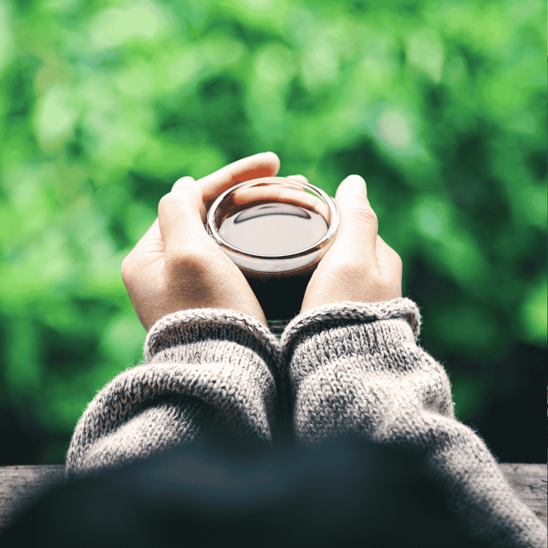 A person wearing a gray sweater holds a cup of coffee or tea with both hands, viewed from above, with a blurred green background suggesting outdoor nature and quiet reflection on new business opportunities.
